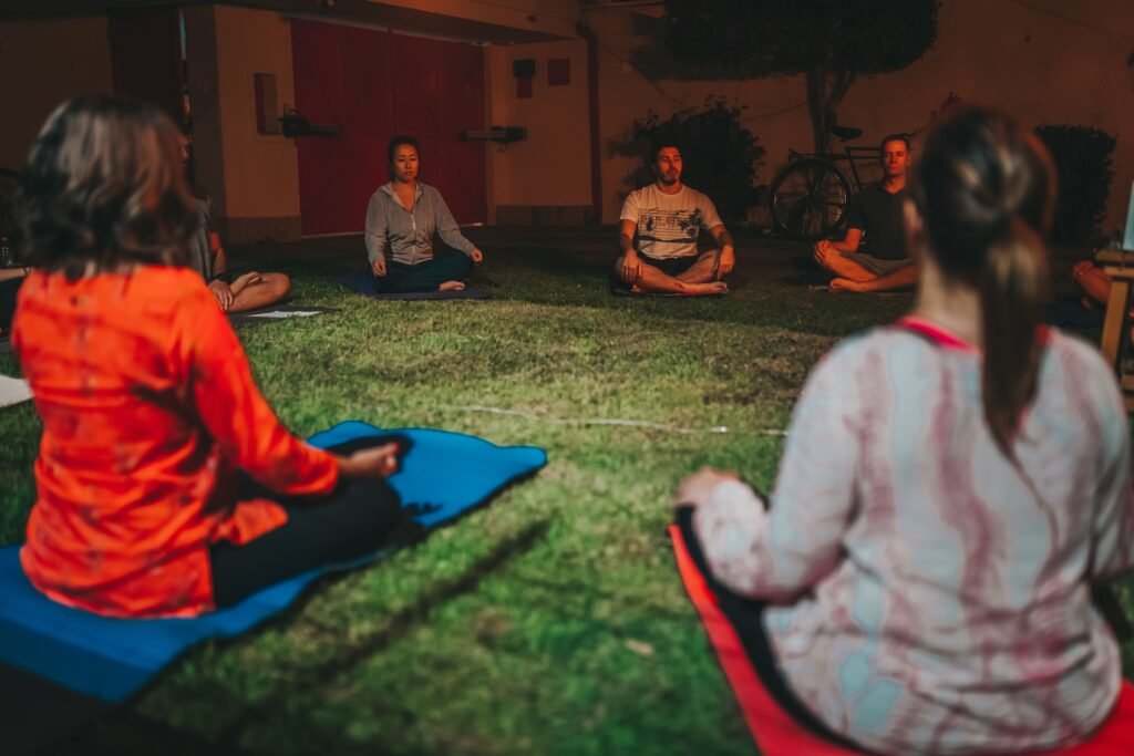 A group meditating cross-legged on grass during a peaceful evening outdoors.